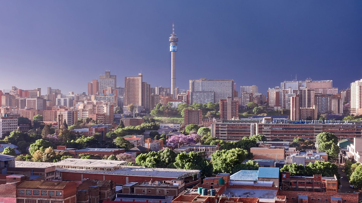 The Johannesburg skyline, with the Hillbrow Tower rising above dense high-rise apartment blocks, jacarandas in bloom and warehouse rooftops in the foreground