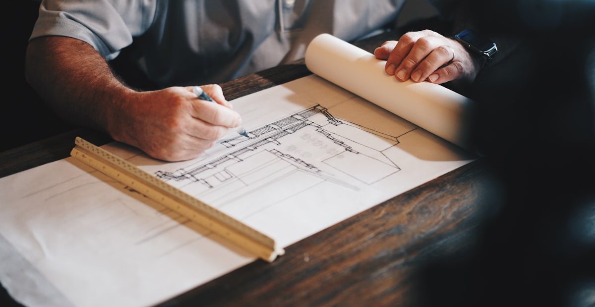 Hands sketching a detailed architectural drawing with a scale ruler on a wooden desk, representing the staged blueprint behind a Decision Intelligence implementation