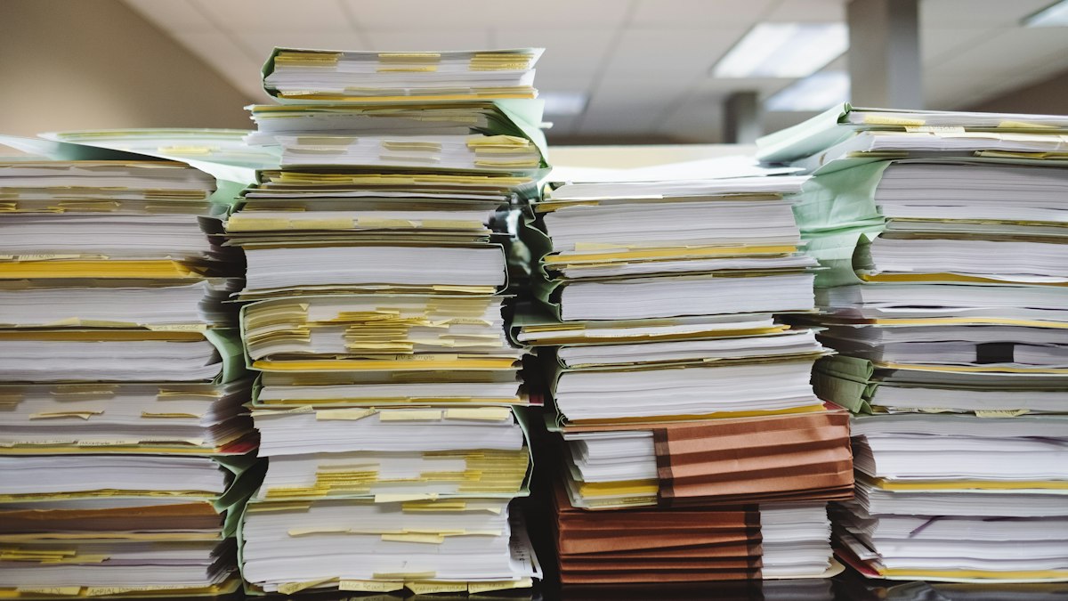 Towering stacks of paper files and folders, bristling with yellow sticky tabs, piled high across desks in an office