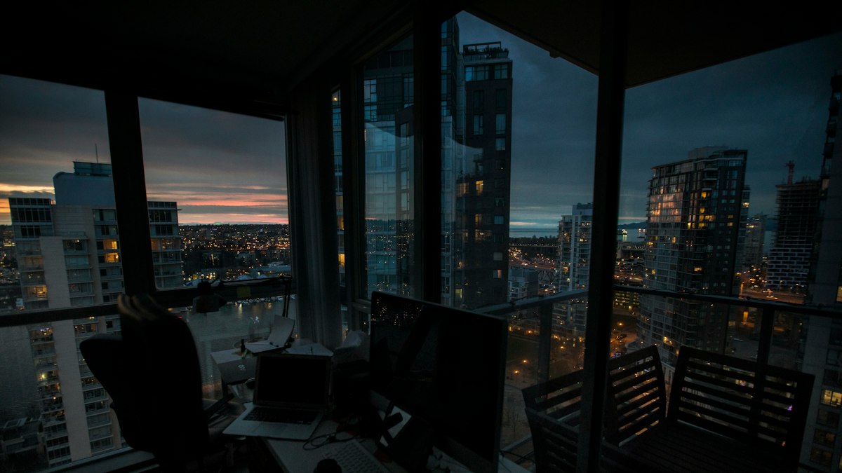 A high-rise office desk with monitors, looking out through floor-to-ceiling glass over a lit city